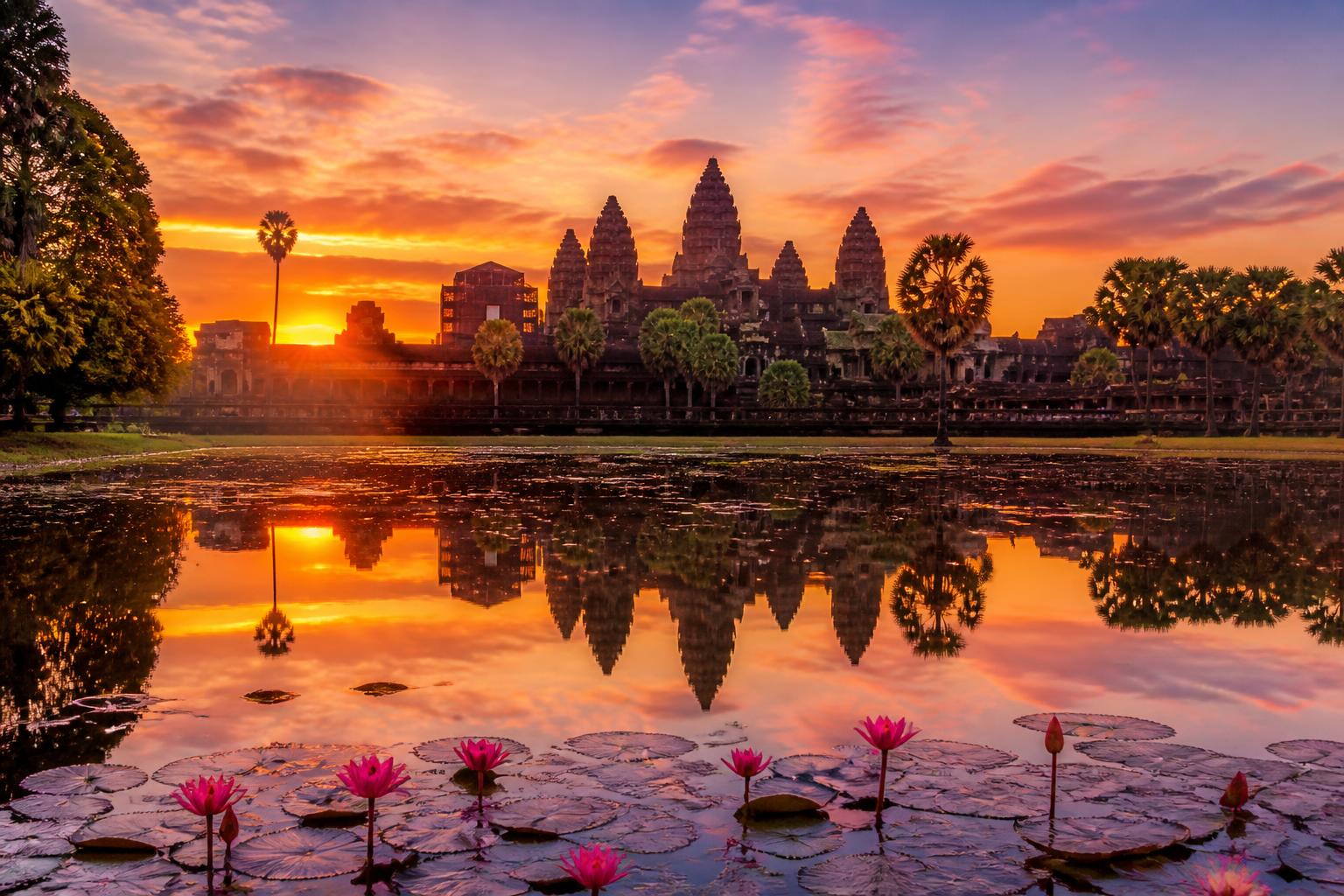 Templos antigos de Angkor Wat em Siem Reap ao nascer do sol, refletidos na água calma com flores de lótus na frente.