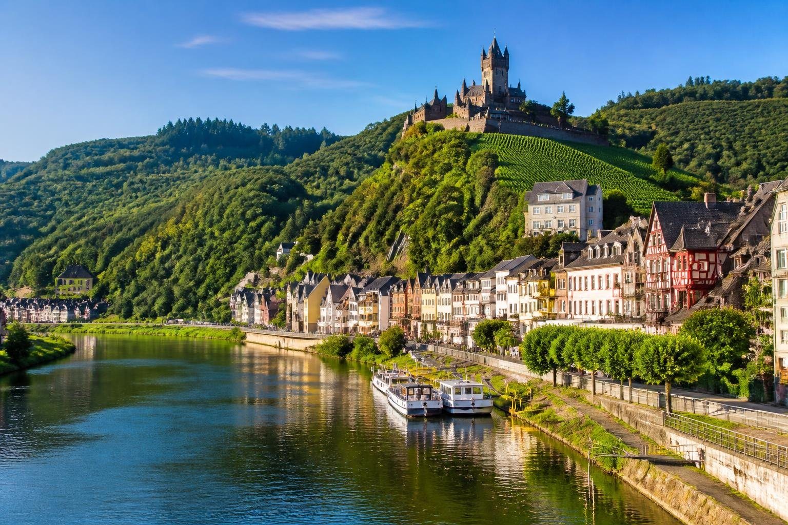 Vista do castelo Reichsburg Cochem no topo da colina, com o rio Mosela e casas tradicionais alemãs ao redor, sob um céu claro e ensolarado.