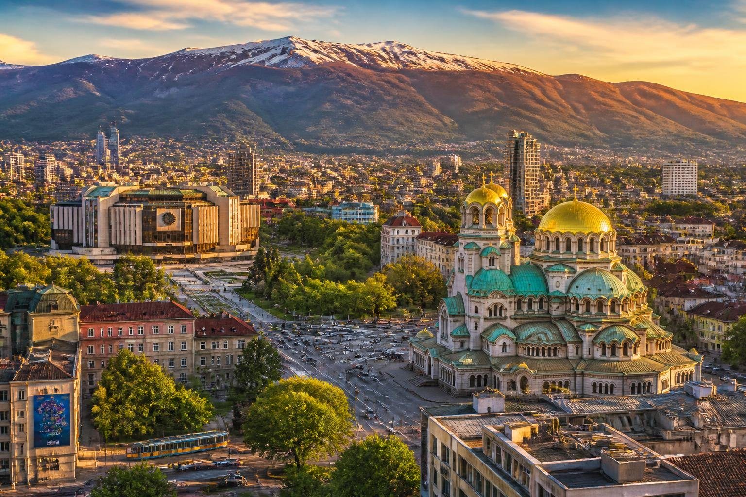 Vista panorâmica da cidade de Sofia, Bulgária, mostrando a Catedral Alexander Nevsky, montanhas ao fundo, prédios históricos e modernos, e áreas verdes.